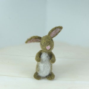 Könnte beinhalten: Eine handgefertigte Hasenfigur aus Filz. Der Hase ist hauptsächlich olivgrün mit einem weißen Bauch und rosa Innenohren und Nase. Der Hase steht auf einer weißen Oberfläche.