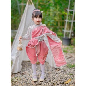May include: A young girl in a traditional Thai costume. The outfit features a pink and white top with intricate lace detailing, paired with matching pink pants. A decorative sash and pearl necklace complete the look. She holds a small woven purse and wears white shoes.