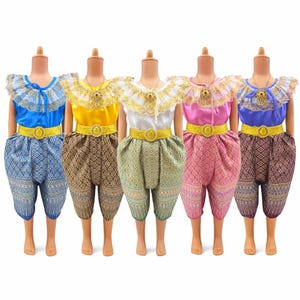 Könnte beinhalten: Fünf traditionelle thailändische Kinderoutfits in Blau, Gelb, Weiß, Rosa und Lila. Jedes Outfit besteht aus einem Oberteil mit Rüschenkragen, einem dekorativen Gürtel und einer gemusterten Hose. Die Outfits werden auf Schaufensterpuppen präsentiert.