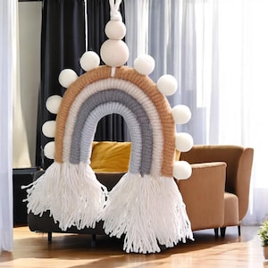 Puede incluir: Un tapiz de pared tejido en forma de arcoíris con flecos blancos y pompones blancos. El arcoíris está hecho de hilo beige, gris y azul claro.