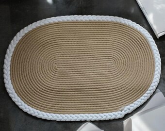 May include: A natural fiber oval rug with a white braided rope border. The rug is made of woven jute and has a natural, earthy tone.