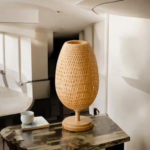 May include: A woven wicker lamp with a natural wood base. The lamp has a round, oval-shaped shade and sits on a wooden table.