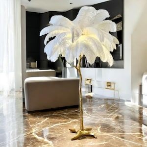 Op de afbeelding: Een witte veren vloerlamp met een gouden metalen voet in de vorm van een tak. De lamp staat in een moderne woonkamer met een witte bank en marmeren vloeren.