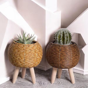 Peut inclure: Deux jardinières rondes en osier avec des pieds en bois. L'une est brun clair et contient une plante succulente, l'autre est brun foncé et contient un cactus. Les jardinières sont sur une surface claire.