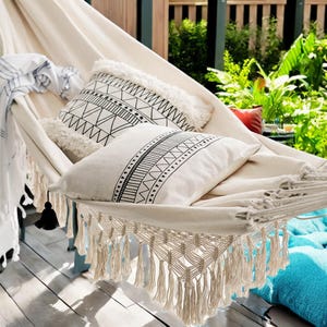 May include: A white hammock with fringe trim and two black and white patterned pillows. The hammock is hanging between two wooden posts.