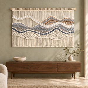 May include: A large rectangular macrame wall hanging featuring a landscape design with wavy layers of cream, tan, light blue, and dark blue knotted cord. The piece is suspended from a horizontal wooden dowel and finished with a long fringe at the bottom.