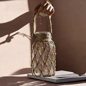 May include: A clear glass jar with a rope handle and a brown rope net covering the jar. The jar is sitting on a white book.