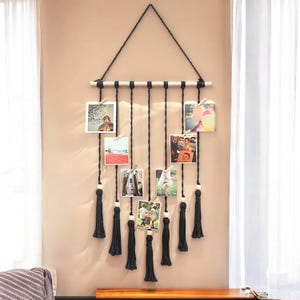 Könnte beinhalten: Ein schwarzer Makramee-Wandbehang mit Holzleiste und schwarzen Quasten. Der Wandbehang hat mehrere Schnüre mit Klammern zum Befestigen von Fotos.