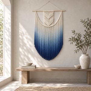 May include: A large macrame wall hanging featuring a central chevron knot pattern that transitions from an off-white top into a deep blue ombre gradient. The piece is suspended from a horizontal wooden dowel with a white cord for mounting.