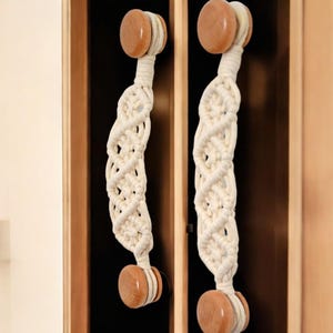 Peut inclure: Deux poignées de tiroir en macramé blanc avec des boutons en bois. Les poignées sont fixées à un meuble en bois.