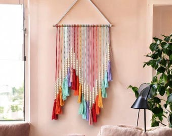 Puede incluir: Un tapiz de macramé de pared colorido con cuentas de madera y borlas. El tapiz está hecho con una variedad de colores, incluyendo rojo, naranja, amarillo, verde, azul y morado.