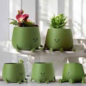 May include: A collection of five green, smiling plant pots with attached legs. Two pots contain plants with flowers and greenery. The pots have a rounded shape and a simple, cartoon-like design, perfect for adding a touch of whimsy to any space.