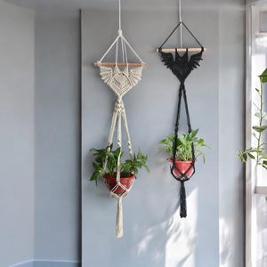 Puede incluir: Dos maceteros colgantes de macramé, uno negro y otro blanco, colgando del techo con plantas en macetas.