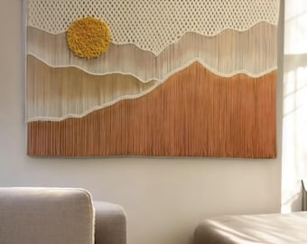 May include: A macrame wall hanging with a yellow sun and white clouds against a brown background.