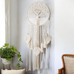 May include: A white macrame dream catcher with a tree of life design and feather accents. The dream catcher is hanging on a white wall.