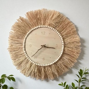 Puede incluir: Un reloj de pared redondo con un diseño bohemio natural. La esfera del reloj es de madera clara con manecillas negras y números sencillos. El reloj está rodeado por un fleco de fibras naturales y un anillo de pequeñas cuentas blancas.