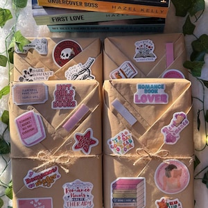 May include: Four brown paper-wrapped books with various romance-themed stickers. The stickers feature phrases like "Romance Book Lover", "Hot Girls Read Books", and "Romance Novels Are My Therapy".