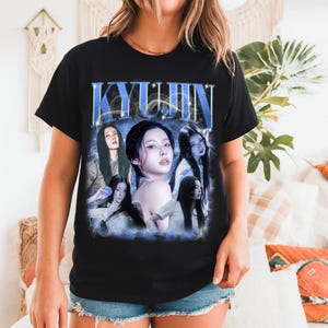 May include: Black t-shirt featuring a graphic of a woman with the name "KYUJIN" in blue text. The graphic includes multiple images of the woman in various poses, surrounded by a blue, smoky effect. The shirt is a classic crew neck.