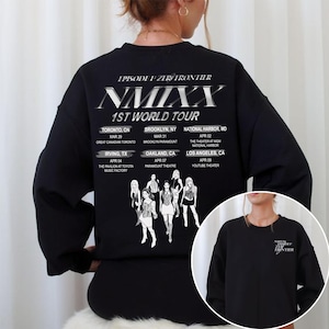 May include: Black sweatshirt featuring the text "NMIXX 1ST WORLD TOUR" with tour dates and locations. The back of the sweatshirt has a graphic of the band members. The sweatshirt is a crew neck style.