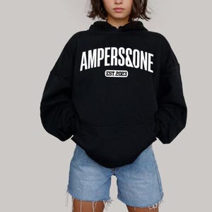 May include: Black hoodie with the text "AMPERS&ONE" in white, with "EST.2023" below. The hoodie has a front pocket and a hood. The person is wearing blue denim shorts.