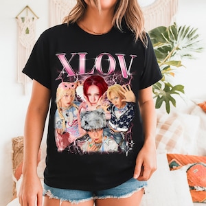 May include: Black t-shirt featuring a graphic of a group of people with the text "XLOV" in pink. The graphic has a vintage, distressed look with lightning bolt accents. The people in the graphic are wearing various outfits.