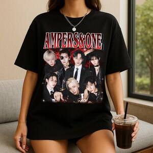 May include: Black t-shirt featuring a group of seven people with the text "AMPERS&ONE" in red and white. The image has a vintage, slightly faded aesthetic. The people are dressed in dark clothing.