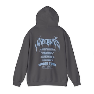 Puede incluir: Sudadera con capucha gris oscuro. La parte trasera presenta texto azul claro: "Stray Kids" en una fuente estilizada, una lista de fechas y lugares de la gira de 2025 y "World Tour 2025".