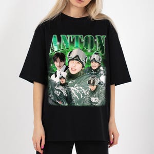 May include: Black t-shirt featuring a graphic design with the name "ANTON" in green, along with multiple images of a person wearing ski goggles and winter clothing. The design has a vintage, distressed look.