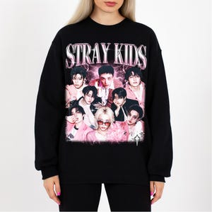 May include: Black sweatshirt featuring the text "STRAY KIDS" in a pink, metallic font above a group photo of nine people. The photo has a pink and purple color scheme, with the people wearing pink and black outfits.