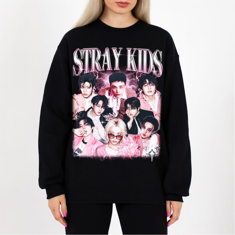 May include: Black sweatshirt featuring the text "STRAY KIDS" in a pink, metallic font above a group photo of nine people. The photo has a pink and purple color scheme, with the people wearing pink and black outfits.
