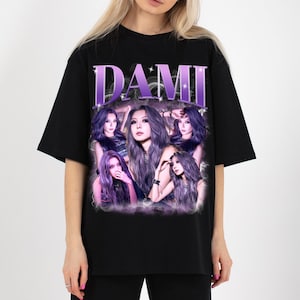 May include: Black t-shirt featuring a graphic of a woman with purple hair and the name "DAMI" in a purple, sparkly font. The graphic includes multiple images of the woman in various poses, with a purple and white lightning effect.