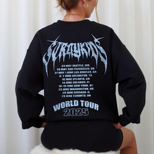 Puede incluir: Sudadera negra con un dise&ntilde;o gr&aacute;fico azul claro en la espalda. El dise&ntilde;o incluye el texto "Stray Kids" en una fuente estilizada, una lista de fechas de la gira y las palabras "World Tour 2025".