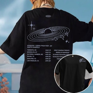 May include: Black t-shirt with a white graphic of a planet and stars, plus the text "EPISODE 1: ZERO FRONTIER" and a tour schedule. The shirt has short sleeves and a crew neck.