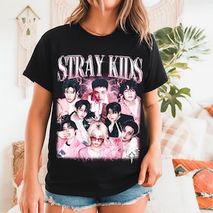 May include: Black t-shirt featuring the text "STRAY KIDS" in a pink and white gradient font above a collage of nine people. The people are wearing various outfits, with pink and white accents.