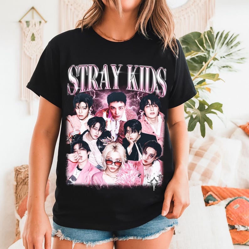May include: Black t-shirt featuring the text "STRAY KIDS" in a pink and white gradient font above a collage of nine people. The people are wearing various outfits, with pink and white accents.