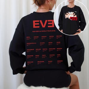 May include: Black sweatshirt featuring the word "EVE" in red block letters, with "2025 BIBI 1st WORLD TOUR [EVE]" printed below. The back of the sweatshirt lists tour locations in red, including Seoul, Chicago, and Los Angeles. A smaller image of the sweatshirt is in the upper right corner.