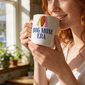 May include: White ceramic mug with the text "DOG MOM ERA" in blue. The mug features a cartoon dog illustration with yellow and pink accents. The mug is held by a person with red hair.