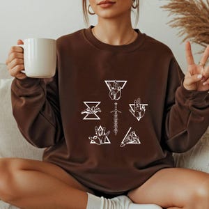 May include: A brown sweatshirt featuring white graphic designs of hands, triangles, and a sword. The sweatshirt has a crew neck and long sleeves. The design is centered on the front of the shirt.