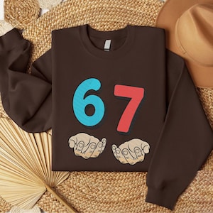 May include: A brown sweatshirt featuring the number "67" in blue and red, with a shadow effect. Below the numbers are two hands. The sweatshirt is displayed on a woven surface with a hat in the background.