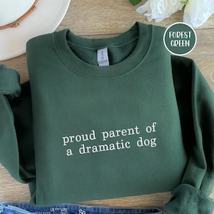 May include: Forest green sweatshirt with the text "proud parent of a dramatic dog" in white. The sweatshirt is folded on a wooden surface, with a white hat and a flower visible in the background.
