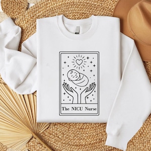 May include: White crew neck sweatshirt with a black and white graphic. The graphic features a baby cradled in hands, with a heart and stars above, and the text "The NICU Nurse" below. The sweatshirt is made of a soft material.