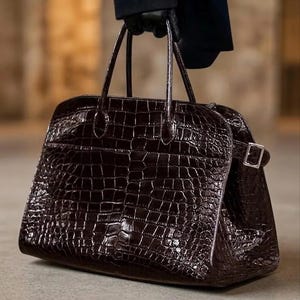 May include: A dark brown, crocodile-textured handbag with two top handles and a rectangular shape. The bag features a silver buckle closure on the side and is held by a person wearing black gloves.