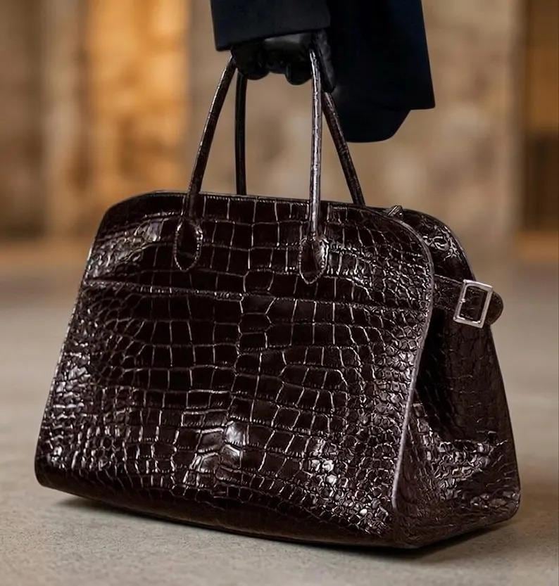 May include: A dark brown, crocodile-textured handbag with two top handles and a rectangular shape. The bag features a silver buckle closure on the side and is held by a person wearing black gloves.