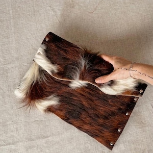 Könnte beinhalten: Rechteckige Clutch aus braun-weißem Kuhfell. Die Clutch hat eine überlappende Klappe, die mit Metallnieten befestigt ist. Das natürliche Fellmuster ist sichtbar, mit einer Mischung aus dunkelbraunen, hellbraunen und weißen Flecken.