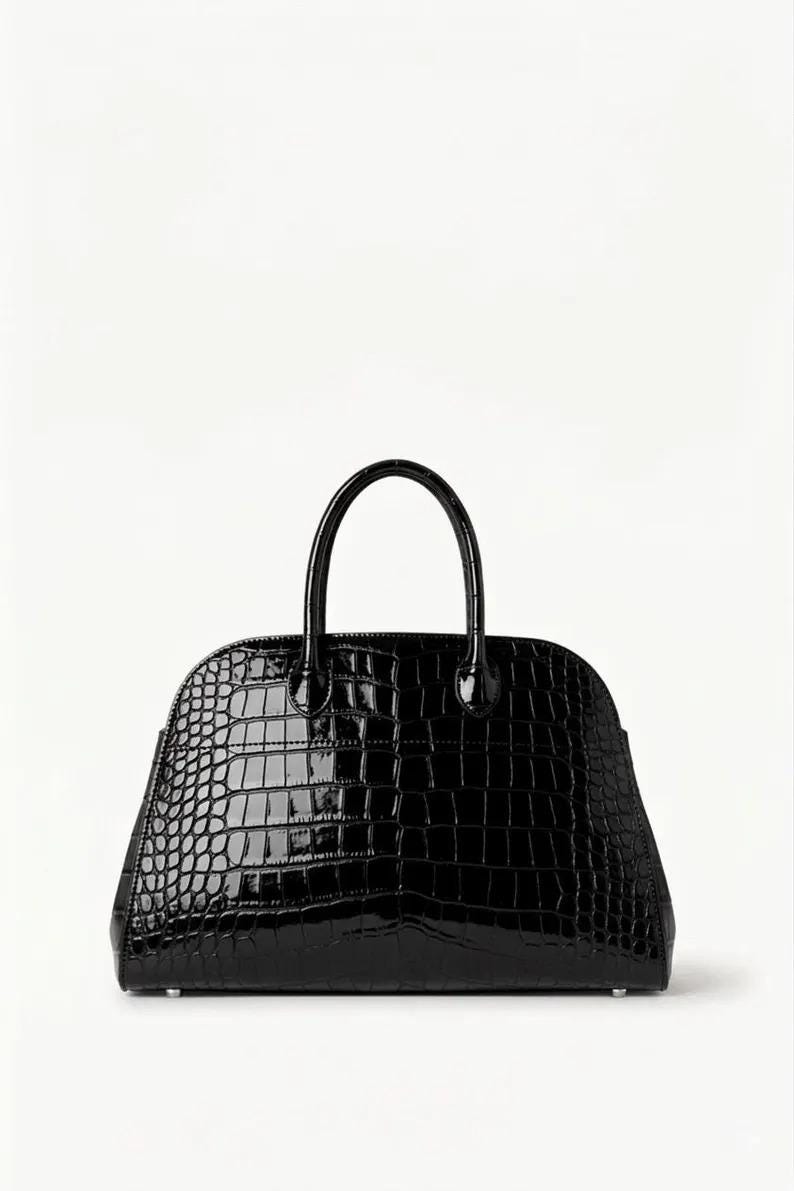 May include: A glossy black handbag with a crocodile skin texture. The bag has a rounded shape with a top handle and a zipper closure. The bag is set against a white background.