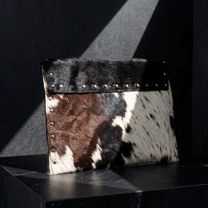 May include: A rectangular clutch bag crafted from cowhide featuring a tri-color pattern of black, brown, and white patches. The bag is accented with a horizontal row of silver-toned metal studs along the top flap and vertical studs along the sides.