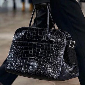 May include: A black, textured leather tote bag with a crocodile skin pattern. The bag has two top handles and a rectangular shape with a buckle closure on the side. The bag is being carried by a person wearing black clothing.