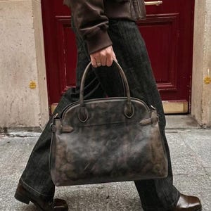 May include: A dark brown leather handbag with a distressed, mottled finish, featuring two rounded top handles, side strap detailing, and a structured silhouette. The bag is held by a person wearing a brown leather jacket and dark denim jeans.