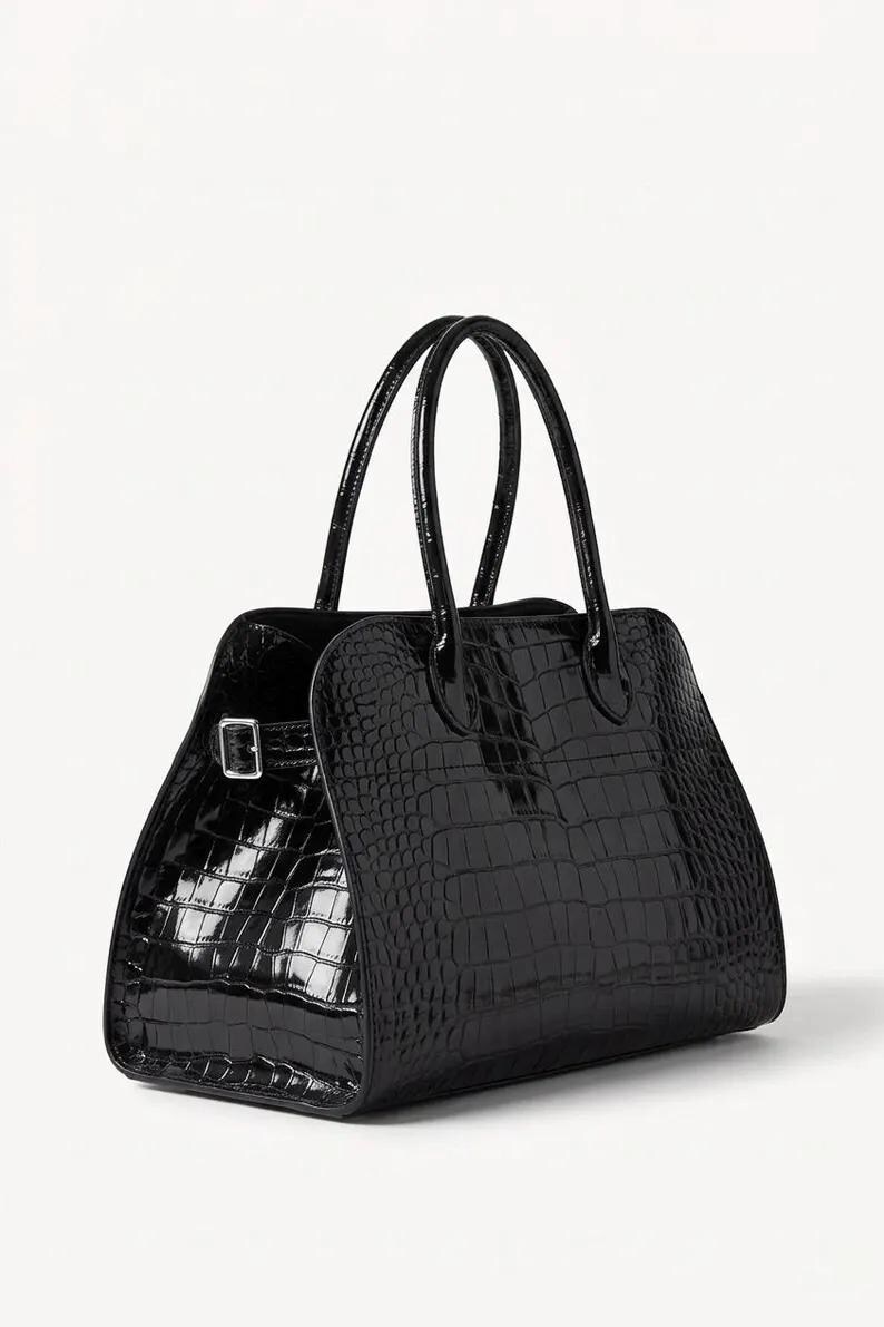 May include: A glossy black handbag with a crocodile skin texture. The bag has two top handles and a rectangular shape with a buckle closure. The bag is set against a white background.
