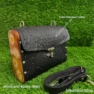 Puede incluir: Bolso bandolera negro de piel grabada con estampado de serpiente con un panel lateral de madera con flores de margarita en resina epoxi. El bolso tiene un cierre plateado, tachuelas metálicas y una correa negra ajustable.